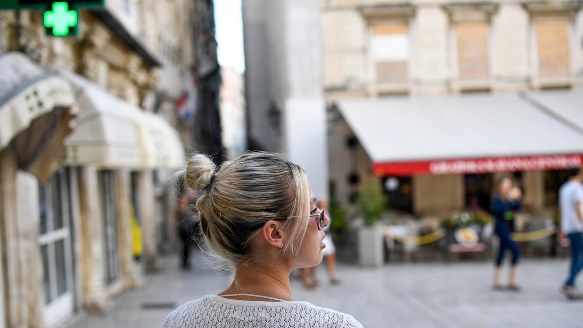 Profile view of a stylish young blonde woman walking through a historic European city square during summer vacation.